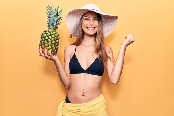 Fiatal szép lány visel bikini és kalap tartja ananász sikoltozó büszke, ünneplő győzelem és a siker nagyon izgatott felemelt karral  - Fotó, kép