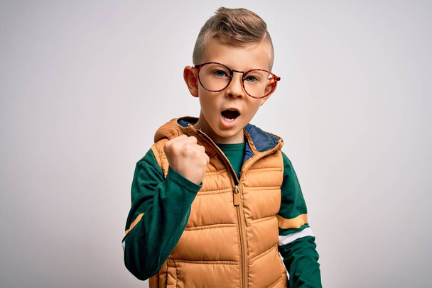 Young little caucasian kid with blue eyes wearing winter coat and smart glasses angry and mad raising fist frustrated and furious while shouting with anger. Rage and aggressive concept. - Photo, Image