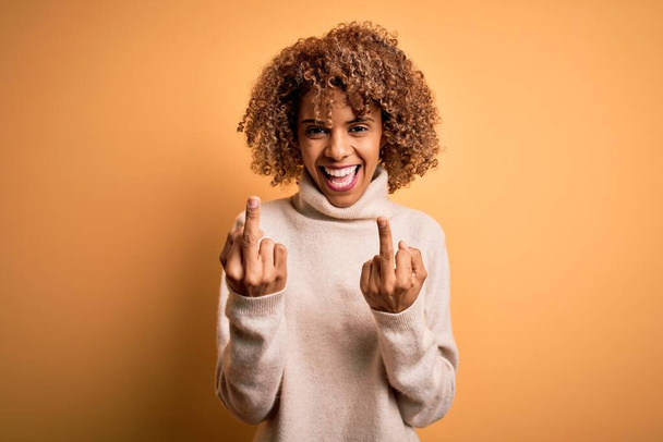 Young beautiful african american woman wearing turtleneck sweater over yellow background Showing middle finger doing fuck you bad expression, provocation and rude attitude. Screaming excited - Foto, Imagen