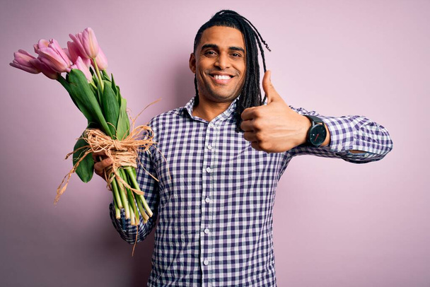 Young african american afro romantic man with dreadlocks holding bouquet of pink tulips happy with big smile doing ok sign, thumb up with fingers, excellent sign - Photo, Image