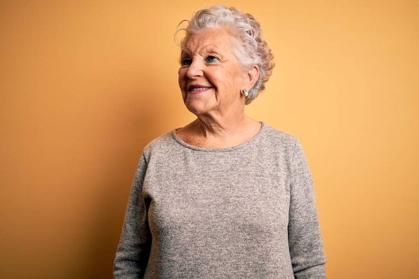 Пожилая красивая женщина в случайной футболке стоит на изолированном желтом фоне, смотрит в сторону с улыбкой на лице, естественным выражением лица. Уверенный смех. - Фото, изображение