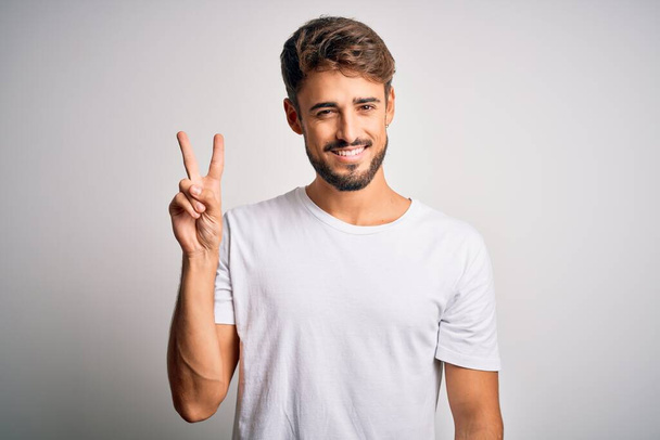 Young handsome man with beard wearing casual t-shirt standing over white background showing and pointing up with fingers number two while smiling confident and happy. - Photo, image