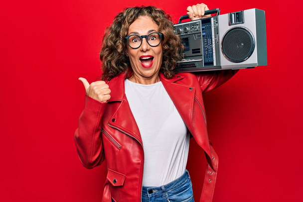 Middle age brunette hipster woman holding retro music boombox over red isolated background pointing thumb up to the side smiling happy with open mouth - Photo, Image