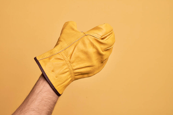 Χέρι του Καυκάσου νεαρός άνδρας με γάντι κηπουρού πάνω από απομονωμένο κίτρινο φόντο κρατώντας αόρατο αντικείμενο, άδειο χέρι κάνει απόκομμα και αρπάζοντας χειρονομία - Φωτογραφία, εικόνα
