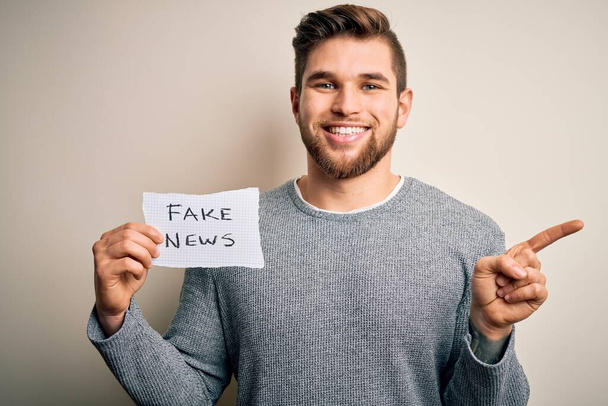 Mladý blonďatý muž s plnovousem a modrýma očima drží papír s falešnou zprávou velmi šťastně ukazuje rukou a prstem na stranu - Fotografie, Obrázek