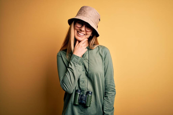 Kaunis blondi tutkimusmatkailija nainen siniset silmät yllään hattu ja lasit kiikarit etsivät luottavainen kamera hymyillen ristissä kädet ja käsi nostetaan leuka. Positiivista.. - Valokuva, kuva