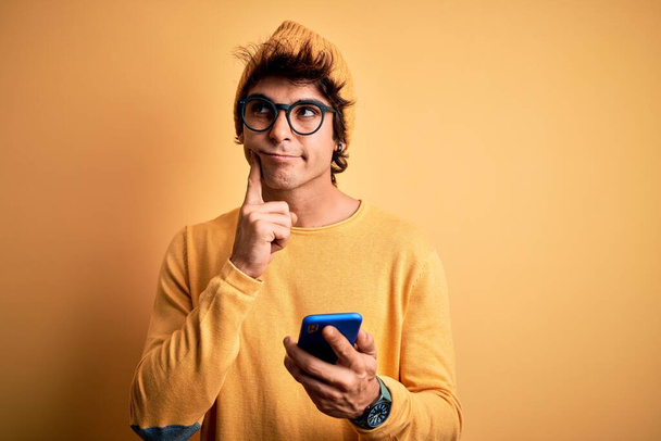 Young handsome man using smartphone wearing glasses over isolated yellow background serious face thinking about question, very confused idea - 写真・画像