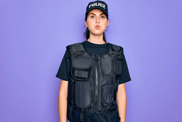 Jonge politievrouw draagt kogelvrije vest uniform over paarse achtergrond puffen wangen met grappig gezicht. Mond opgeblazen met lucht, gekke uitdrukking. - Foto, afbeelding