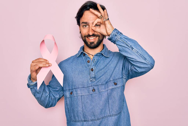 Jeune homme hispanique tenant un ruban rose de sensibilisation au cancer du sein avec le visage souriant et un signe ok avec la main sur l'oeil en regardant à travers les doigts - Photo, image