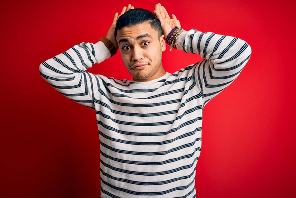Young brazilian man wearing casual striped t-shirt standing over isolated red background Doing bunny ears gesture with hands palms looking cynical and skeptical. Easter rabbit concept. - Photo, Image