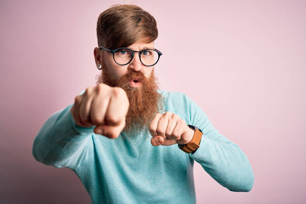 Beau homme à tête rouge irlandais portant une barbe et des lunettes sur fond rose et isolé Piqûre de poing à combattre, attaque agressive et en colère, menace et violence - Photo, image