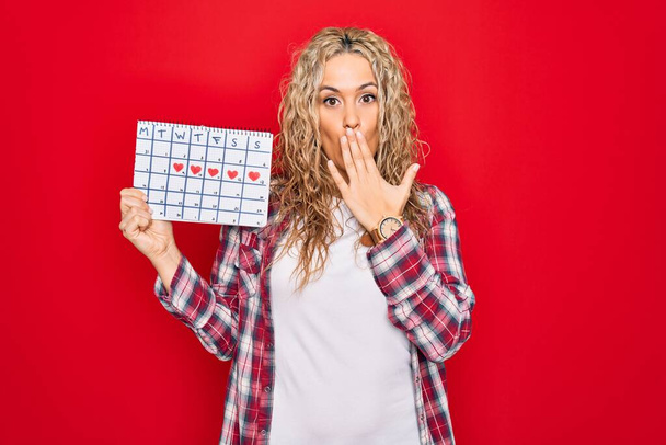 Junge schöne blonde Frau mit einem Kalender, der den Menstruationszyklus kontrolliert und den Mund mit der Hand bedeckt, schockiert und ängstlich vor Fehlern. Überraschte Miene - Foto, Bild