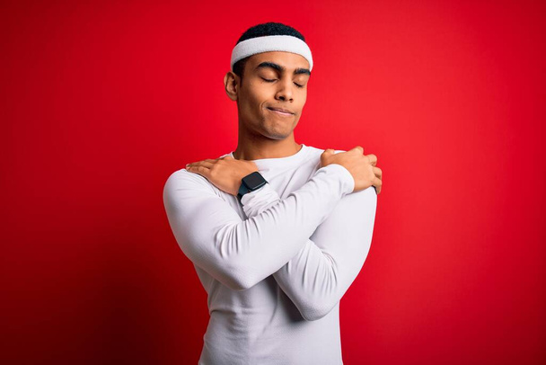 Junger gut aussehender afrikanisch-amerikanischer Sportler, der Sportbekleidung über rotem Hintergrund trägt und sich glücklich und positiv umarmt, selbstbewusst lächelt. Selbstliebe und Selbstfürsorge - Foto, Bild