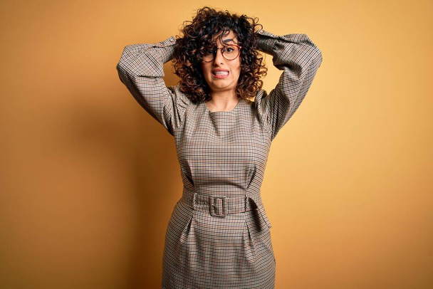 Beautiful arab business woman wearing dress and glasses standing over yellow background Crazy and scared with hands on head, afraid and surprised of shock with open mouth - Photo, Image