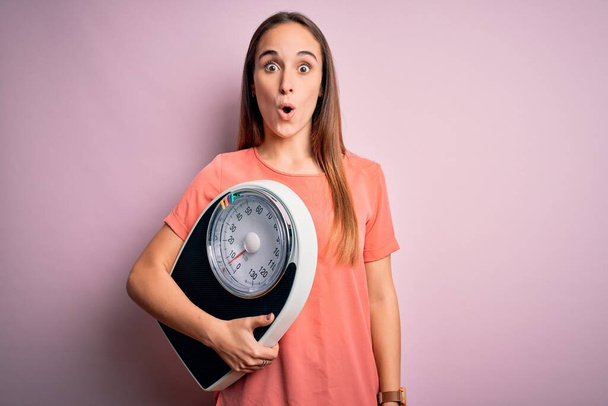 Young beautiful woman controlling weight holding weighting machine over pink background scared in shock with a surprise face, afraid and excited with fear expression - Photo, Image