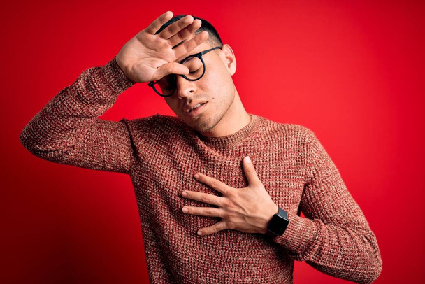 Jeune homme hispanique portant des verres de nerf sur fond rouge Touchant le front pour maladie et fièvre, grippe et rhume, maladie virale - Photo, image