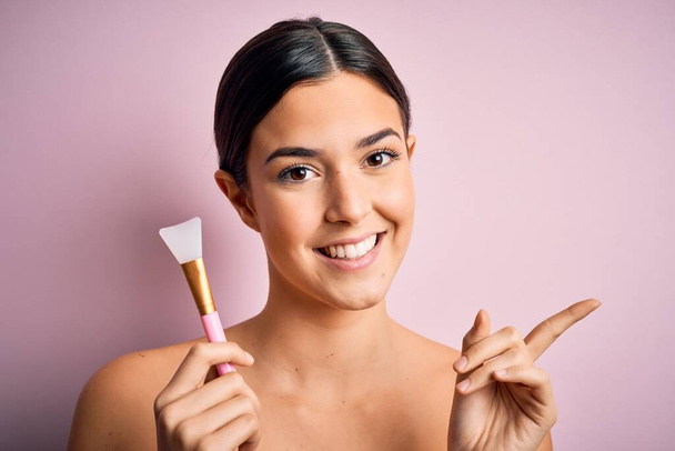 Junge schöne Mädchen Anwendung von Kosmetik mit Spachtel über rosa Hintergrund sehr glücklich zeigt mit der Hand und dem Finger zur Seite - Foto, Bild