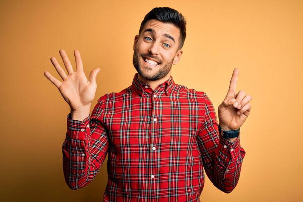 Jonge knappe man dragen casual shirt staande over geïsoleerde gele achtergrond tonen en wijzend met de vingers nummer zes terwijl glimlachend zelfverzekerd en gelukkig. - Foto, afbeelding