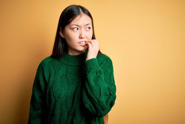 Young beautiful asian woman wearing green winter sweater over yellow isolated background looking stressed and nervous with hands on mouth biting nails. Anxiety problem. - Foto, imagen
