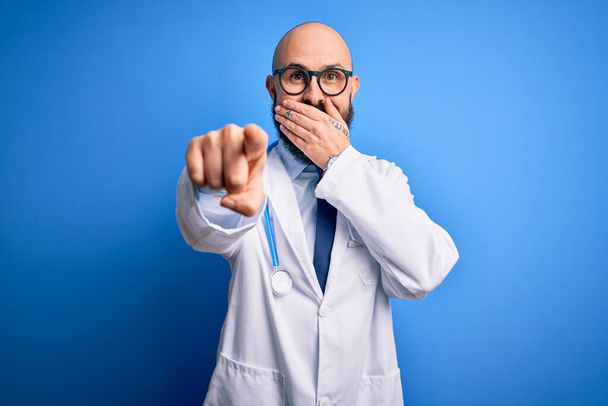 Komea kalju lääkäri, jolla on parta silmälasit ja stetoskooppi sinisen taustan päällä, nauraa sinulle, osoittaa sormella kameraa käsin suun yli, häpeä ilme - Valokuva, kuva