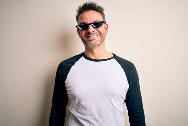 Jonge knappe man draagt grappige schurk leven zonnebril meme over witte achtergrond met een vrolijke en koele glimlach op het gezicht. Geluksvogel. - Foto, afbeelding