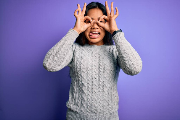 Young beautiful asian girl wearing casual sweater standing over isolated purple background doing ok gesture like binoculars sticking tongue out, eyes looking through fingers. Crazy expression. - Fotografie, Obrázek