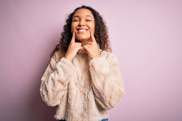 Young beautiful woman with curly hair wearing casual sweater standing over pink background Smiling with open mouth, fingers pointing and forcing cheerful smile - Photo, Image
