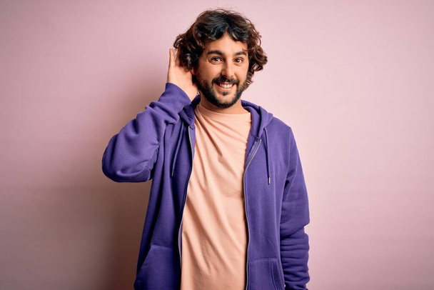 Young handsome sporty man with beard wearing casual sweatshirt over pink background smiling with hand over ear listening an hearing to rumor or gossip. Deafness concept. - Photo, Image