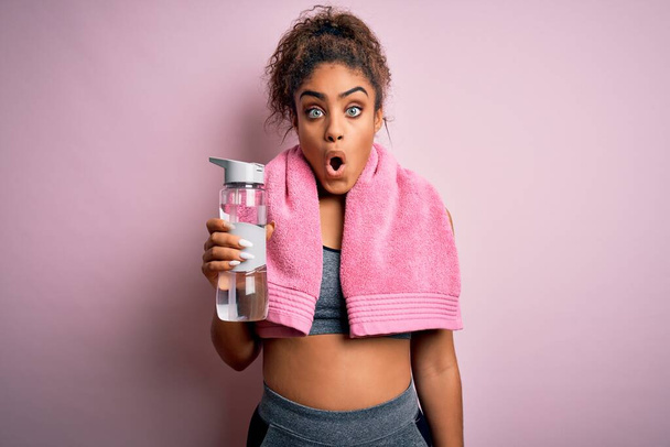 Jeune fille sportive afro-américaine faisant du sport portant une serviette de toilette buvant une bouteille d'eau effrayée en état de choc avec un visage surprise, effrayée et excitée par l'expression de la peur
 - Photo, image