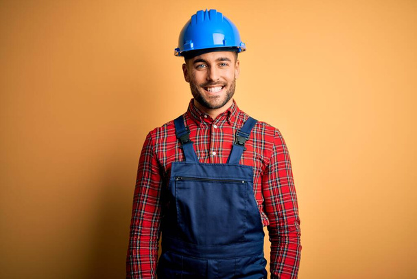 Молодой строитель в строительной форме и шлеме безопасности на жёлтом фоне со счастливой и прохладной улыбкой на лице. Счастливчик
. - Фото, изображение