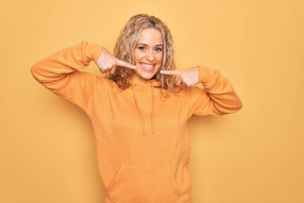 Sarışın, güzel ve sportif bir kadın sarı arka planda rahat bir tişört giyiyor. Gülümsüyor, parmaklarını ve ağzını göstererek gülümsüyor. Diş sağlığı konsepti. - Fotoğraf, Görsel
