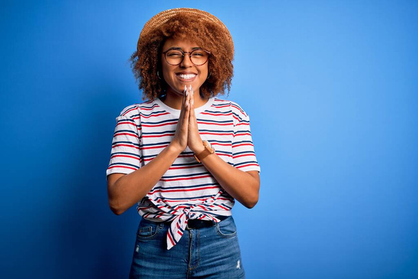 Afroamerikanerin mit lockigem Haar im Urlaub mit Sommermütze und gestreiftem T-Shirt, die mit den Händen zusammen betet und um Vergebung bittet, lächelt zuversichtlich. - Foto, Bild