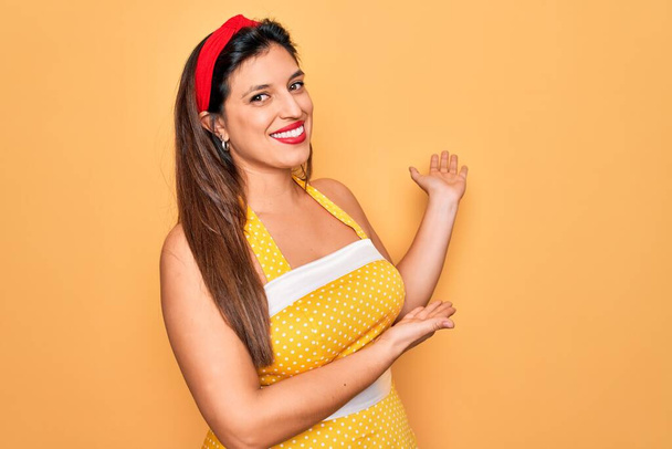 Sarı arka planda 50 'li yaşlarda seksi giyinen Latin asıllı genç bir kadın gülümseyerek içeri girmeye davet ediyor. - Fotoğraf, Görsel
