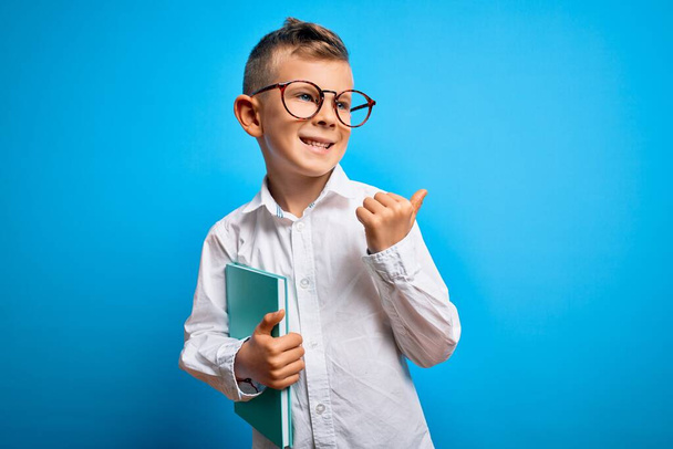 Young little caucasian student kid wearing smart glasses and holding a book from school pointing and showing with thumb up to the side with happy face smiling - Photo, Image