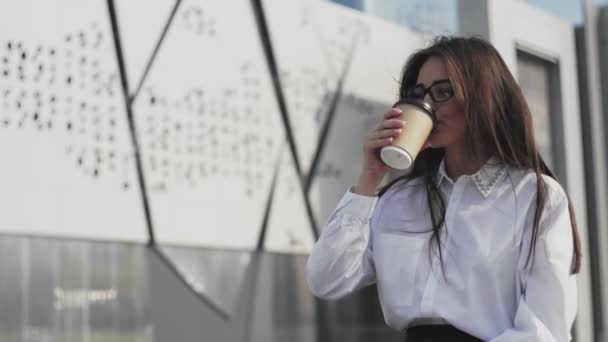 Gyönyörű üzletasszony szemüvegben, okostelefonon beszél és kávét iszik. Modern épület háttérrel - Felvétel, videó