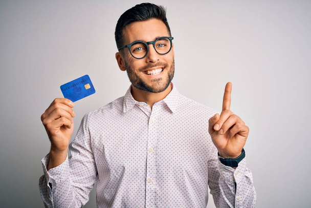Genç bir iş adamı kredi kartını soyutlanmış arka planda tutarken bir fikre ya da soru işaretine şaşırır ve mutlu yüzle işaret eder. - Fotoğraf, Görsel