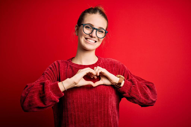 Jonge mooie roodharige vrouw draagt casual trui over geïsoleerde rode achtergrond glimlachend in liefde doen hart symbool vorm met de handen. Romantisch concept. - Foto, afbeelding