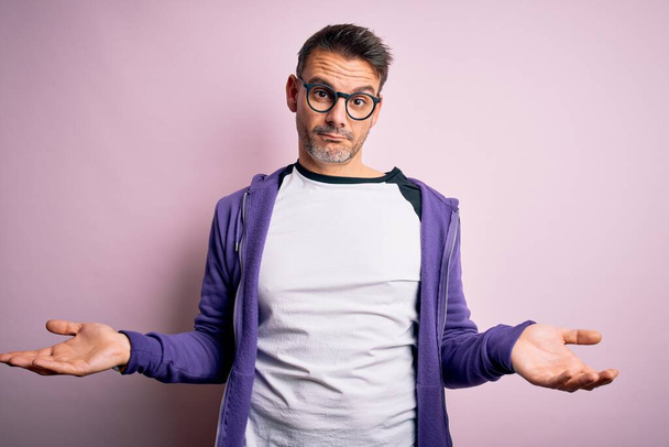Jeune bel homme portant un sweat-shirt violet et des lunettes debout sur fond rose sans idée et confus avec les bras ouverts, aucune idée concept
. - Photo, image
