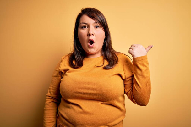 Beautiful brunette plus size woman wearing casual sweater over isolated yellow background Surprised pointing with hand finger to the side, open mouth amazed expression. - Фото, изображение