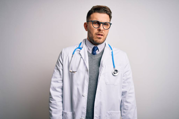 Young doctor man with blue eyes wearing medical coat and stethoscope over isolated background In shock face, looking skeptical and sarcastic, surprised with open mouth - Foto, immagini