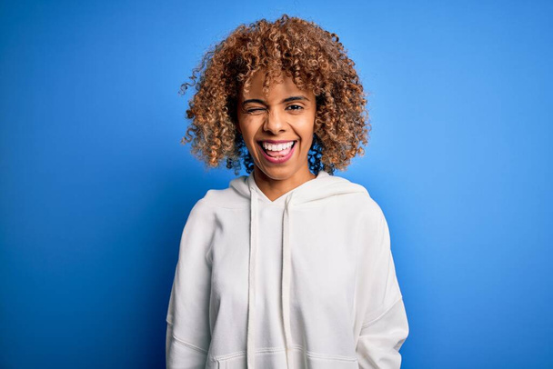Beautiful african american sporty woman wearing casual sweatshirt over blue background winking looking at the camera with sexy expression, cheerful and happy face. - Foto, Imagem