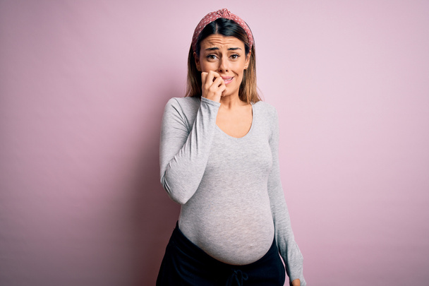 Fiatal gyönyörű barna nő terhes vár baba felett elszigetelt rózsaszín háttér keres stresszes és ideges a kezét a száj harapdáló körmök. Szorongás. - Fotó, kép