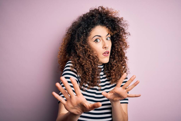 Young beautiful woman with curly hair and piercing wearing casual striped t-shirt afraid and terrified with fear expression stop gesture with hands, shouting in shock. Panic concept. - Фото, изображение