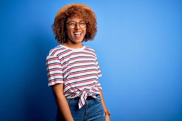 African American woman with curly hair on vacation wearing summer hat and striped t-shirt winking looking at the camera with sexy expression, cheerful and happy face. - Photo, Image