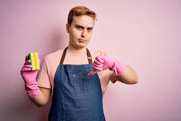 Knappe roodharige man doet huishoudelijk werk met schort en handschoenen met schonere geurstof met boos gezicht, negatief teken toont een hekel aan duimen naar beneden, afwijzing concept - Foto, afbeelding