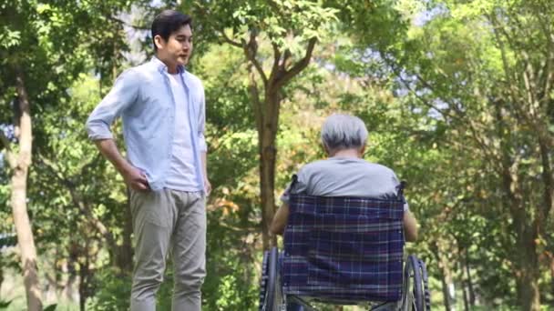 young asian adult son talking to wheelchair bound demoralized senior father outdoors in a park - Footage, Video