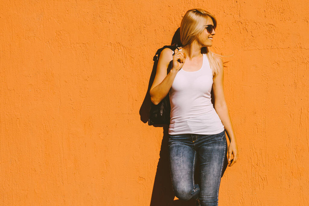 young  smiling blonde woman in white tank top and sunglasses against yellow wall outdoor in city summer heat  - Foto, Imagen