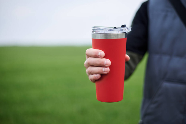 Close-up picture of male hand, holding red travel mug with hot tea coffee drink outside in the field in spring summer. Healthy lifestyle concept. - Photo, Image