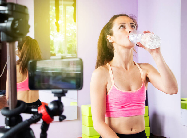A video of a blog, movie or video tutorial. The girl drinks water after intense training, sports and yoga. The phone is on a tripod and shoots a blogger - Foto, imagen