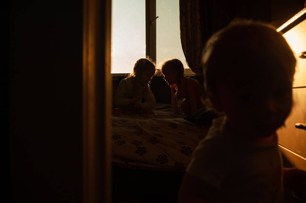 gyerekek játszanak a szobában a lenyugvó nap sugaraiban. Kisgyerekek otthon a háttérben. A nap kisüt az ablakon a gyerekek hátterére. Sziluett felvétel gyermekekről otthon a lenyugvó nap sugaraiban. - Fotó, kép